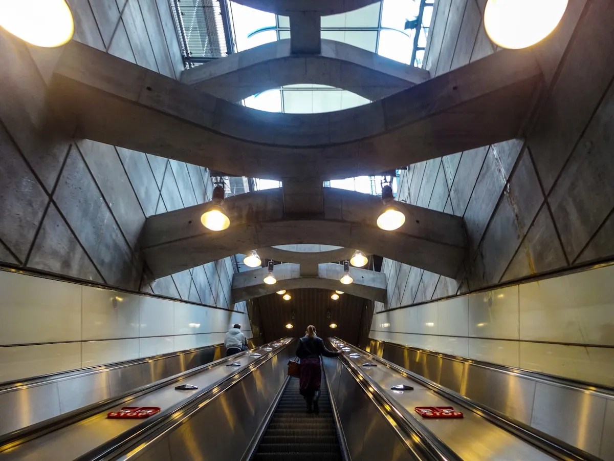 Passengers ride long escalators up through a deep concrete cavern in a modern metro station, with geometric structural beams overhead